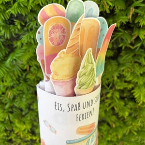 May include: A cylindrical paper container featuring a light blue ice cream truck illustration and the text "EIS, SPA&szlig; UND SCH&Ouml;NE FERIEN!" filled with various die-cut paper ice cream treats including popsicles, cones, and soft serve.
