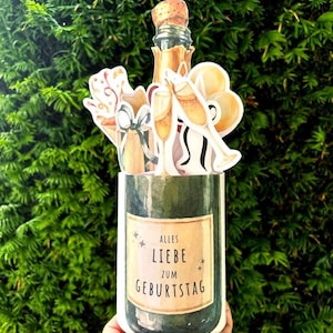 Peut inclure: Une carte de vœux en trois dimensions avec une base en forme de bouteille de champagne vert foncé et une étiquette beige portant l'inscription ALLES LIEBE ZUM GEBURTSTAG. Le haut présente des illustrations de bouteille, flûtes et cadeaux.