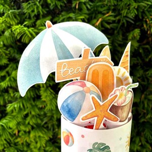 May include: A cylindrical container decorated with beach-themed icons including a beach ball, a monstera leaf, and sunglasses. The top features cutouts of a blue and white umbrella, a wooden beach sign, an orange popsicle, and a coconut drink.