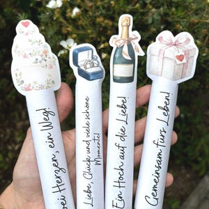 May include: Four white cylindrical paper scrolls featuring watercolor illustrations at the top: a tiered cake, a ring box, a champagne bottle, and a gift box. Each scroll displays German text: Zwei Herzen, ein Weg; Liebe, Gl&uuml;ck und viele sch&ouml;ne Momente; Ein Hoch auf die Liebe; Gemeinsam f&uuml;rs Leben!