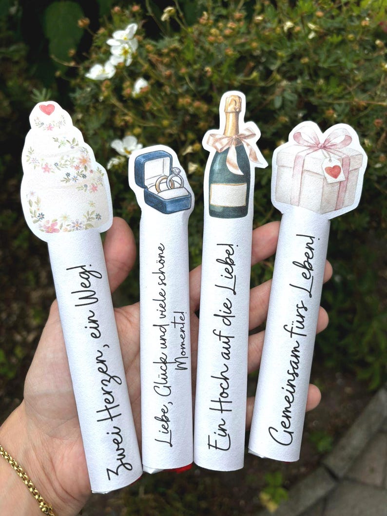 May include: Four white cylindrical paper scrolls featuring watercolor illustrations at the top: a tiered cake, a ring box, a champagne bottle, and a gift box. Each scroll displays German text: Zwei Herzen, ein Weg; Liebe, Gl&uuml;ck und viele sch&ouml;ne Momente; Ein Hoch auf die Liebe; Gemeinsam f&uuml;rs Leben!