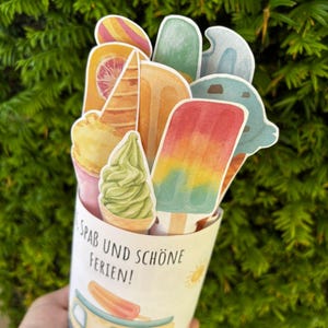 May include: A cylindrical paper container featuring a colorful illustration of an ice cream truck and the text "SPAB UND SCH&Ouml;NE FERIEN!". The container is filled with various paper cutouts of ice cream cones, popsicles, and soft serve treats.