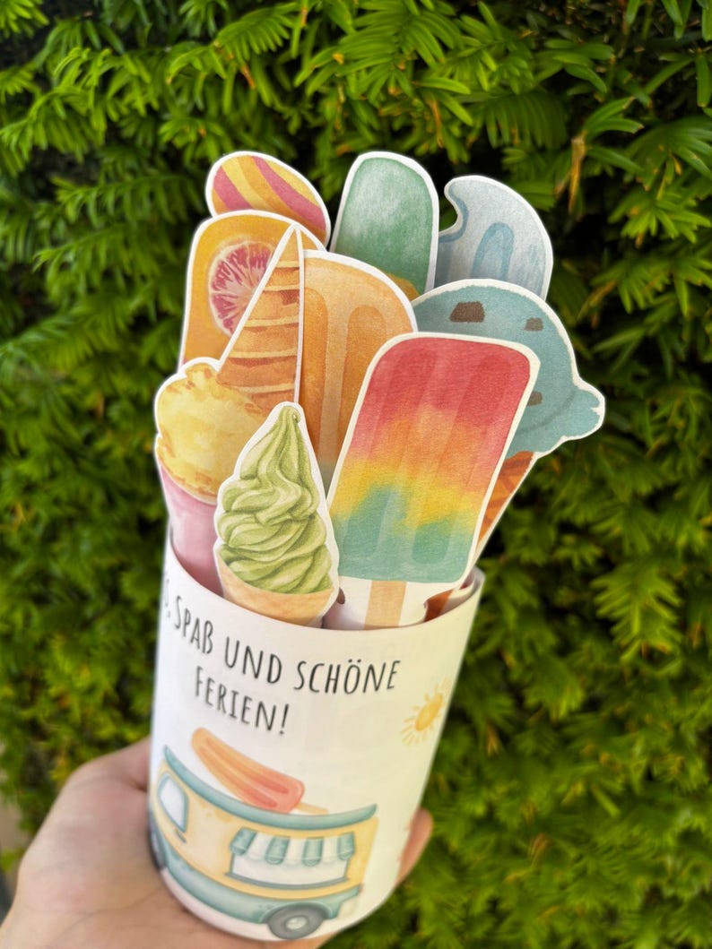 May include: A cylindrical paper container featuring a colorful illustration of an ice cream truck and the text "SPAB UND SCH&Ouml;NE FERIEN!". The container is filled with various paper cutouts of ice cream cones, popsicles, and soft serve treats.