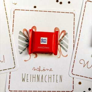 Op de afbeelding: Een kleine rode Ritter Sport Marzipan chocoladereep op een witte kaart met een gedrukte illustratie van een strik en de tekst 'sch&ouml;ne WEIHNACHTEN'. De kaart heeft een bruine stippellijnrand en is omringd door kleine gouden sterconfetti.