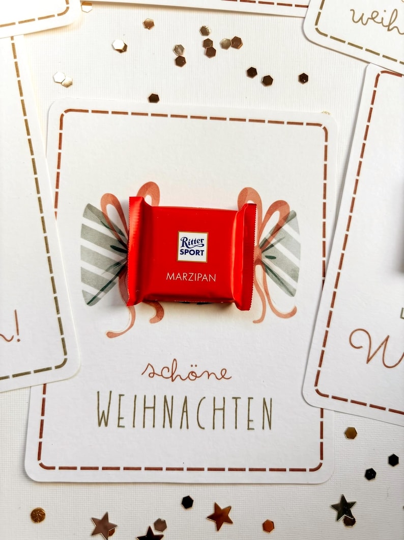 Op de afbeelding: Een kleine rode Ritter Sport Marzipan chocoladereep op een witte kaart met een gedrukte illustratie van een strik en de tekst 'sch&ouml;ne WEIHNACHTEN'. De kaart heeft een bruine stippellijnrand en is omringd door kleine gouden sterconfetti.