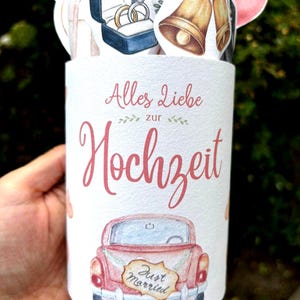 May include: A cylindrical white container featuring the German text Alles Liebe zur Hochzeit, a pink vintage car with a Just Married sign, and decorative cutouts of gold wedding bells, a blue ring box with two rings, and pink balloons.