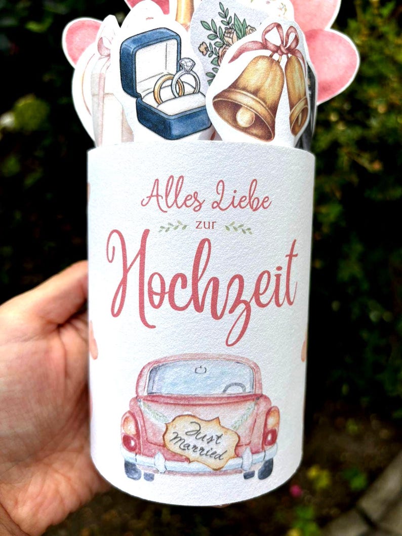 May include: A cylindrical white container featuring the German text Alles Liebe zur Hochzeit, a pink vintage car with a Just Married sign, and decorative cutouts of gold wedding bells, a blue ring box with two rings, and pink balloons.