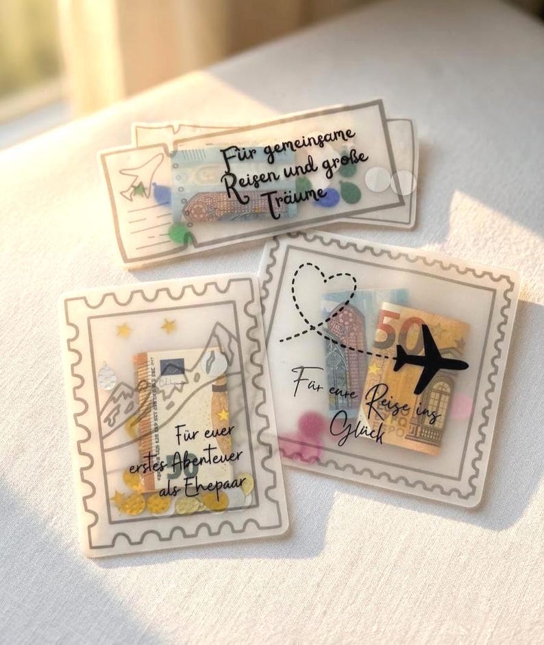 May include: Three translucent, rectangular envelopes designed like postage stamps and tickets, containing folded Euro banknotes and colorful confetti. The envelopes feature black printed text in German, including phrases like F&uuml;r gemeinsame Reisen und gro&szlig;e Tr&auml;ume, F&uuml;r euer erstes Abenteuer als Ehepaar, and F&uuml;r eure Reise ins Gl&uuml;ck.