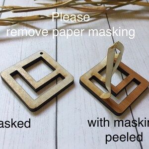 May include: Two wooden squares with a geometric design. The square on the left has a paper masking covering the design. The square on the right has the paper masking peeled off, revealing the natural wood underneath. The text "Please remove paper masking" is written above the squares. The text "masked" is written below the square on the left. The text "with masking peeled" is written below the square on the right.