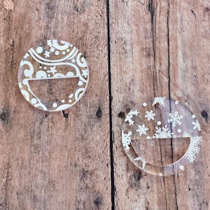 May include: Two clear acrylic ornaments with white designs. One ornament has a snowflake pattern and the other has a swirling pattern. Both ornaments have a cut-out section in the middle.