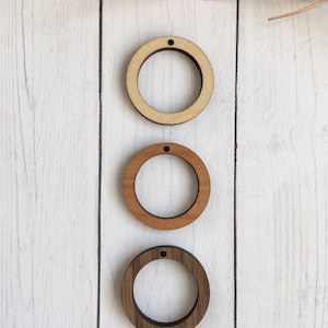 May include: Three wooden circle cutouts in varying shades of brown, from light to dark. Each circle has a small hole at the top, suggesting they are designed for hanging or crafting. The cutouts are arranged vertically against a white wood-grain background.
