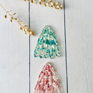 May include: Two clear plastic hair clips with a unique, wavy, tree-like design. One clip is decorated with teal glitter and the other with pink glitter.