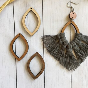 May include: Three wooden earring blanks in the shape of a teardrop with a hole at the top. The blanks are light brown and have a natural wood finish. A gray macrame earring with a wooden bead is also shown.