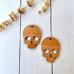 May include: Two unfinished wooden skull shapes, each with a hole at the top for hanging. The skulls are on a white wooden background.