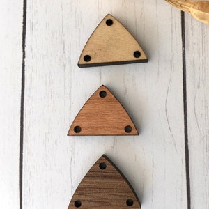 May include: Three wooden triangle-shaped beads in varying shades of brown. Each bead has three small holes at the corners and one at the top. The beads are arranged vertically against a white wood-grain background.