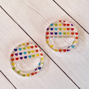 May include: Two clear acrylic round ornaments with a rainbow heart pattern. The ornaments have a cut-out section in the bottom half.