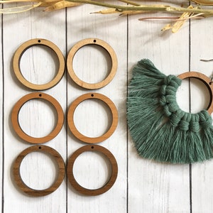 May include: Six unfinished wooden circles with holes in the top, arranged in two rows of three. The circles are different sizes and are on a white wooden background. A green tassel is in the upper right corner of the image.