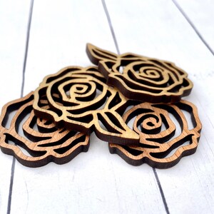 May include: Three wooden rose cutouts, each with a detailed, intricate design. The roses are stacked on top of each other, with the bottom rose partially visible. The roses are a light brown color.