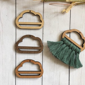 May include: Three wooden cloud-shaped ornaments with a hole at the top for hanging. Each cloud has a cutout in the middle. The clouds are arranged vertically on a white wooden surface. A green tassel hangs from the bottom of the rightmost cloud.