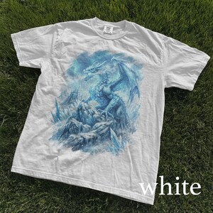 May include: White t-shirt featuring a detailed blue dragon illustration. The dragon is perched on a rocky landscape with icy crystals, set against a cloudy sky. The word "white" is printed at the bottom.