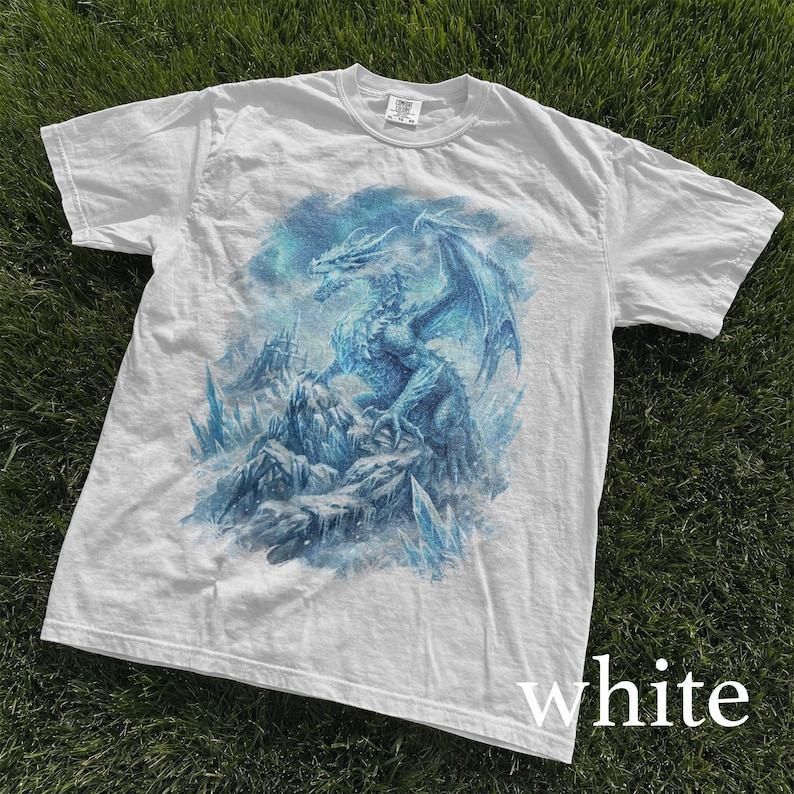 May include: White t-shirt featuring a detailed blue dragon illustration. The dragon is perched on a rocky landscape with icy crystals, set against a cloudy sky. The word "white" is printed at the bottom.