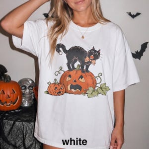 May include: A white t-shirt featuring a graphic print of a black cat with an orange polka dot bow tie standing on a large carved jack-o'-lantern, accompanied by a smaller jack-o'-lantern and green leaves. The word white is printed at the bottom.