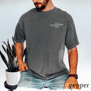 May include: Dark gray t-shirt with the text "Cool Dads Club" in white script. The shirt is a short-sleeved, crew neck style. The model is wearing blue jeans and a brown watch.