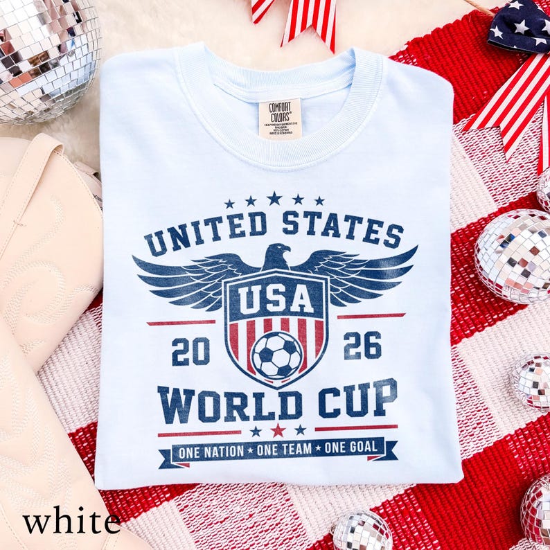 May include: A light blue crew neck t-shirt featuring a dark blue and red graphic print that reads UNITED STATES 2026 WORLD CUP with an eagle, a shield containing a soccer ball, and the text ONE NATION ONE TEAM ONE GOAL at the bottom.