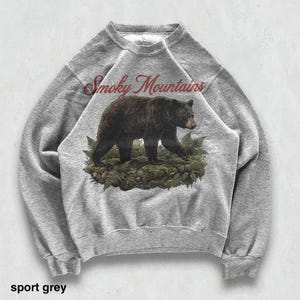 May include: Sport grey sweatshirt featuring a black bear walking on green foliage. The words "Smoky Mountains" are arched above the bear in a burgundy script. The sweatshirt is a crew neck style.