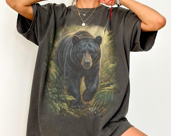 May include: Dark grey t-shirt with a detailed illustration of a black bear in a forest. The bear is the central image, surrounded by green foliage. The word "pepper" is printed in white at the bottom left of the t-shirt.