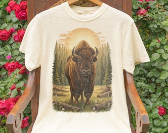 May include: Ivory-coloured t-shirt with a bison illustration set in a natural landscape. The design includes a sunset, trees, and fallen logs. The shirt is displayed on a wooden hanger.