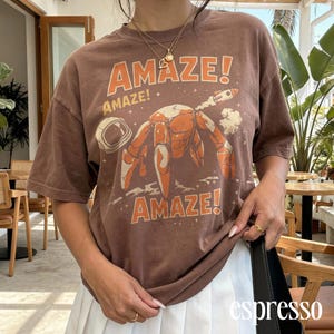 May include: Brown t-shirt with the word "AMAZE!" in orange, along with a graphic of a crab in space. The shirt has short sleeves and a crew neck. The word "espresso" is printed on the bottom right.