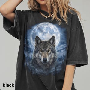 May include: Black t-shirt featuring a wolf with yellow eyes, set against a full moon and forest backdrop. The design is printed on a dark, oversized tee. The word "black" is printed at the bottom left.