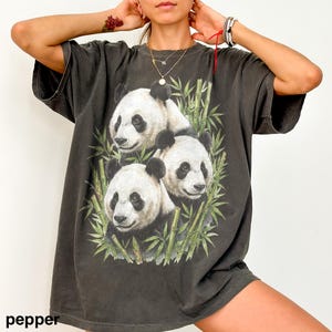 May include: Dark gray t-shirt featuring a graphic of three pandas surrounded by bamboo. The pandas have black and white fur. The word "pepper" is printed at the bottom left of the shirt.