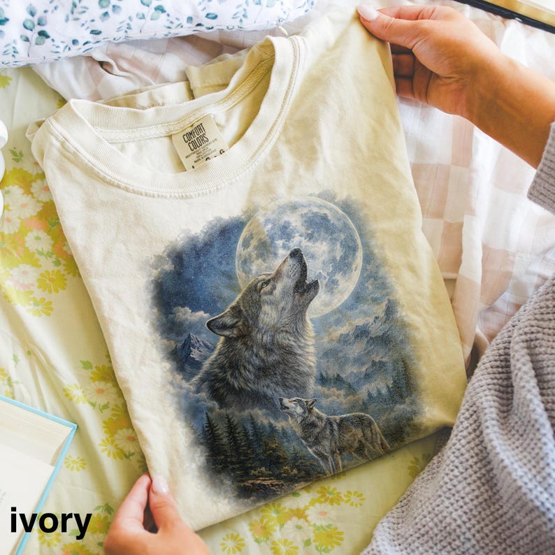 May include: Ivory-colored t-shirt featuring a detailed graphic of a howling wolf under a full moon. The design includes a second wolf and a forest scene. The shirt is made of a soft material and has a classic crew neck.