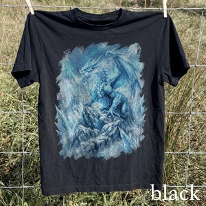 May include: Black t-shirt featuring a detailed blue and white dragon illustration. The dragon is depicted with large wings and is set against a backdrop of ice and snow. The word "black" is printed in white at the bottom.