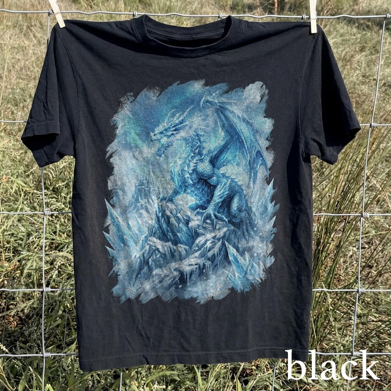 May include: Black t-shirt featuring a detailed blue and white dragon illustration. The dragon is depicted with large wings and is set against a backdrop of ice and snow. The word "black" is printed in white at the bottom.