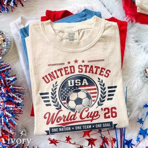 May include: A cream-colored t-shirt featuring a graphic with the text UNITED STATES USA World Cup '26 and ONE NATION ONE TEAM ONE GOAL. The design includes a soccer ball, a United States flag motif, laurel branches, and red and blue stars.