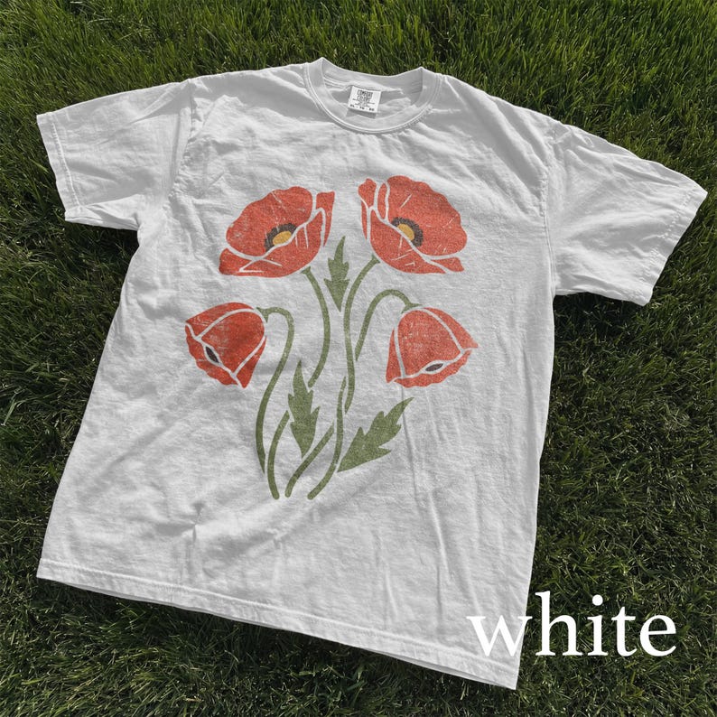 Op de afbeelding: Wit T-shirt met een bloemenontwerp van vier rode klaprozen en groene stelen en bladeren. Het woord "white" is in het wit op de onderkant van het shirt gedrukt. Het shirt is gemaakt van een zacht, comfortabel materiaal.