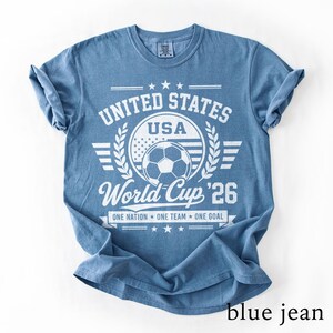 May include: A blue short-sleeve t-shirt featuring a white graphic print that reads UNITED STATES USA World Cup '26 ONE NATION ONE TEAM ONE GOAL with a soccer ball and stars. The shirt has a crew neckline and a distressed, vintage-style design.