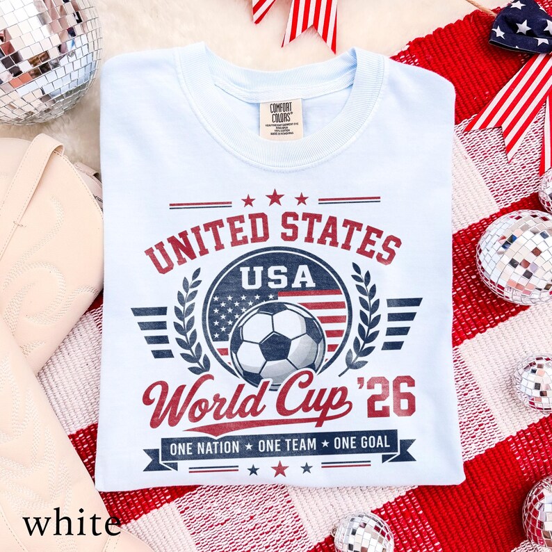 May include: A light blue t-shirt featuring a graphic with the text UNITED STATES USA World Cup '26 and ONE NATION ONE TEAM ONE GOAL. The design includes a soccer ball, a United States flag motif, stars, and laurel branches in red and navy blue.