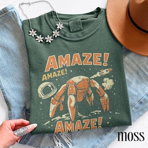 May include: Moss green t-shirt with the word "AMAZE!" in orange, repeated three times, and a cartoon crab with a rocket ship and astronaut helmet graphic. The shirt is displayed with a necklace, hat, and jeans.