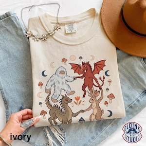 May include: An ivory t-shirt featuring a graphic print of a white yeti, a red dragon, a brown jackalope, and a grey creature holding hands in a circle. The design includes small mushrooms, moons, and a flower. The shirt has a crew neckline.