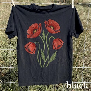 Op de afbeelding: Zwart T-shirt met een bloemenontwerp van vier rode klaprozen met groene stelen en bladeren. Het woord "black" staat rechtsonder in het wit gedrukt. Het shirt hangt aan een waslijn.