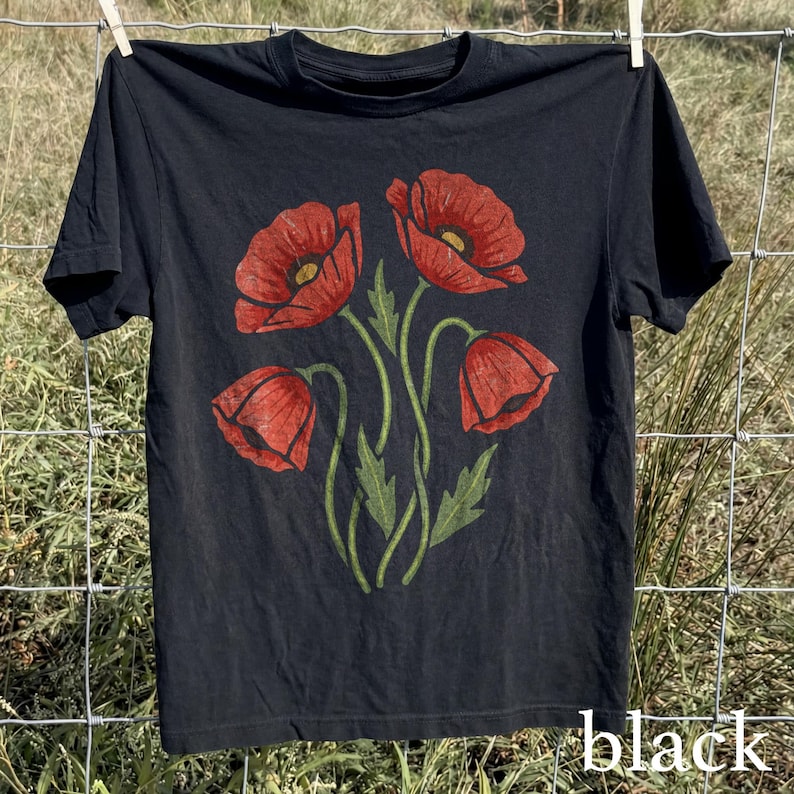 Op de afbeelding: Zwart T-shirt met een bloemenontwerp van vier rode klaprozen met groene stelen en bladeren. Het woord "black" staat rechtsonder in het wit gedrukt. Het shirt hangt aan een waslijn.