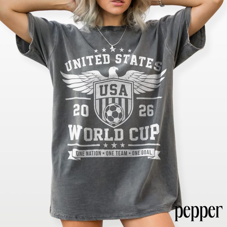 May include: A charcoal gray short-sleeve t-shirt featuring a white graphic print that reads UNITED STATES 2026 WORLD CUP with an eagle emblem, a soccer ball, and the text ONE NATION ONE TEAM ONE GOAL displayed on the front of the garment.