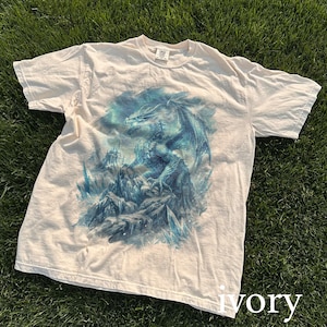 May include: Ivory-colored t-shirt featuring a detailed illustration of a blue dragon perched on a rocky landscape with icy formations. The dragon has outstretched wings and is set against a cloudy background. The word "ivory" is printed at the bottom.