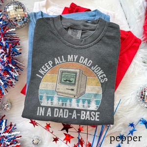 Op de afbeelding: Donkergrijs T-shirt met de tekst "I KEEP ALL MY DAD JOKES IN A DAD-A-BASE" en een retro computer graphic. Het shirt is opgevouwen bovenop andere opgevouwen shirts in rood, wit en blauw. Het shirt is gemaakt door Comfort Colors.