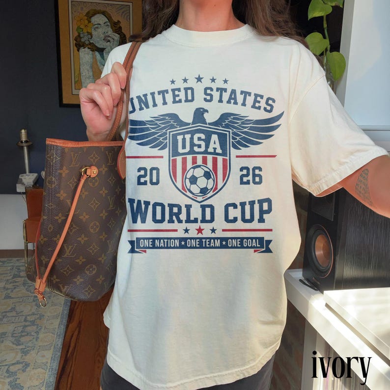 May include: An ivory t-shirt featuring a blue and red graphic print that reads UNITED STATES 2026 WORLD CUP, with an eagle emblem, a soccer ball, and the text ONE NATION ONE TEAM ONE GOAL. The shirt is paired with a brown monogrammed tote bag.