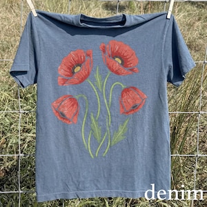 Op de afbeelding: Een denim blauw t-shirt met een bloemenontwerp van rode klaprozen met groene stelen en bladeren. Het woord "denim" staat in het wit rechtsonder gedrukt. Het shirt hangt aan een waslijn.